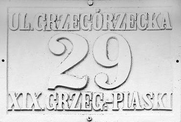 Fot. 27. W roku 1881 Rada Miasta Krakowa, utrzymując dotychczasowy podział na 8 dzielnic katastralnych (jedynie Dzielnicę I w miejsce nazwy „Kraków” nazwano „Śródmieściem”), wprowadziła nowe zasady ponumerowania orientacyjnego domów w mieście. Zasady te przetrwały do dzisiaj: numeracja domów ulicami (a nie dzielnicami katastralnymi, jak poprzednio), początek numerowania od strony bliższej Rynkowi Głównemu (na Stradomiu bliższej ul. Stradomskiej, na Kazimierzu bliższej ul. Krakowskiej), numery nieparzyste po prawej stronie. Wprowadzono jednolite dla całego miasta kolory tabliczek z nazwami ulic i tabliczek orientacyjnych z numerami domów: litery i cyfry koloru czarnego, tło białe, obwódki tablic niebieskie (DzRMK 1881, nr 1, s. 2–3). Przydzielone wówczas nowe numery domów pozostały już z reguły niezmienione do naszych czasów (zmiany w przynależności dzielnicowej ulic nie miały znaczenia dla numeru domu), natomiast sam wzór tabliczek wprawdzie był aktualny jeszcze w okresie okupacji, jednak już w okresie międzywojennym podlegał wymianie na znany nam co do kolorystyki z codzienności wzór nowy, z białymi literami i cyframi na niebieskim tle, z białą obwódką. Powyżej: 4) tabliczka już z okresu Wielkiego Miasta Krakowa (Grzegórzki–Piaski przyłączono do Krakowa w 1910 r., ustanawiając dla tej dawnej gminy wiejskiej odrębną dzielnicę katastralną miasta o numerze XIX).