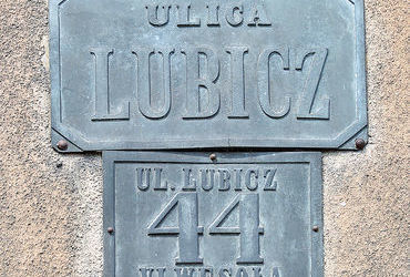 Fot. 28. W roku 1881 Rada Miasta Krakowa, utrzymując dotychczasowy podział na 8 dzielnic katastralnych (jedynie Dzielnicę I w miejsce nazwy „Kraków” nazwano „Śródmieściem”), wprowadziła nowe zasady ponumerowania orientacyjnego domów w mieście. Zasady te przetrwały do dzisiaj: numeracja domów ulicami (a nie dzielnicami katastralnymi, jak poprzednio), początek numerowania od strony bliższej Rynkowi Głównemu (na Stradomiu bliższej ul. Stradomskiej, na Kazimierzu bliższej ul. Krakowskiej), numery nieparzyste po prawej stronie. Wprowadzono jednolite dla całego miasta kolory tabliczek z nazwami ulic i tabliczek orientacyjnych z numerami domów: litery i cyfry koloru czarnego, tło białe, obwódki tablic niebieskie (DzRMK 1881, nr 1, s. 2–3). Przydzielone wówczas nowe numery domów pozostały już z reguły niezmienione do naszych czasów (zmiany w przynależności dzielnicowej ulic nie miały znaczenia dla numeru domu), natomiast sam wzór tabliczek wprawdzie był aktualny jeszcze w okresie okupacji, jednak już w okresie międzywojennym podlegał wymianie na znany nam co do kolorystyki z codzienności wzór nowy, z białymi literami i cyframi na niebieskim tle, z białą obwódką. Powyżej: 2) zachowana i służąca do dzisiaj para tabliczek: z nazwą ulicy i z numerem domu.